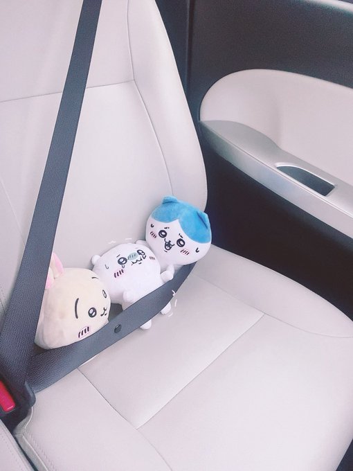 Three small plush toys (Chiikawa-style characters including one with cat ears and a blue-hooded figure) are wedged into the seatbelt of a white leather car seat.