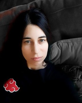 A portrait photo of a young woman with dark hair and fair skin, wearing a dark top, with a small red cloud icon (resembling the Akatsuki symbol from Naruto) in the bottom left corner.