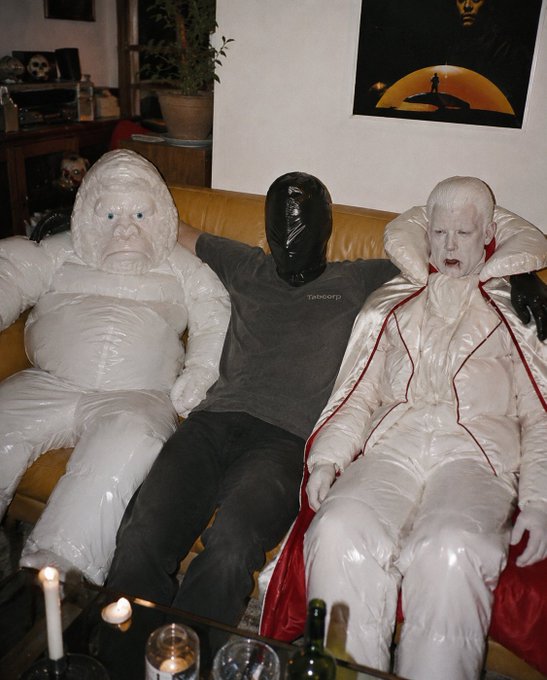 A person wearing a black latex mask sits on a yellow couch between two figures dressed in elaborate white costumes—one resembling a puffy snowman-like creature and the other a pale vampire-like character with a red-lined cape.