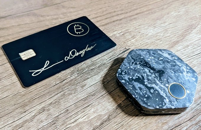 A sleek black Bitcoin card signed by "Douglas" sits next to a hexagonal Bitkey hardware wallet featuring a marbled gray finish, resting on a wooden surface—showcasing the complementary tools discussed in the post about joining the Bitcoin ecosystem with Bitkey and CashApp.