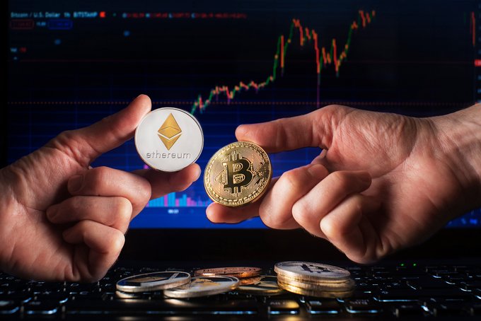 Two hands hold physical coins representing Ethereum (left) and Bitcoin (right) against a backdrop of a stock chart on a computer screen, with additional cryptocurrency coins scattered on a keyboard.