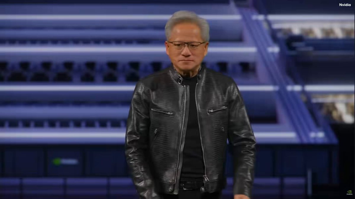 The image features a man wearing a black leather jacket, standing behind a blue screen displaying text that reads "Jensen Huang calls $NBIS and $CRWV word class AI clouds Nebius: •  $27B AI cloud deal with Meta : ...: Multi billion dollar cloud deal with Anthropic :  ...: 5GW Partnership with Nvidia". The man is wearing glasses, and he appears to be talking in front of the screen.