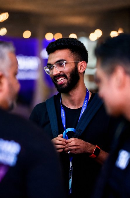 A young bearded man wearing glasses and a lanyard smiles during a conversation at what appears to be a tech conference or event.