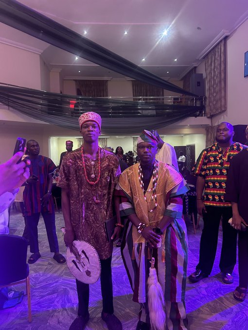 Two young men wearing traditional African attire, including coral beads and patterned robes with caps, stand together at an indoor event with purple lighting and a balcony in the background.
