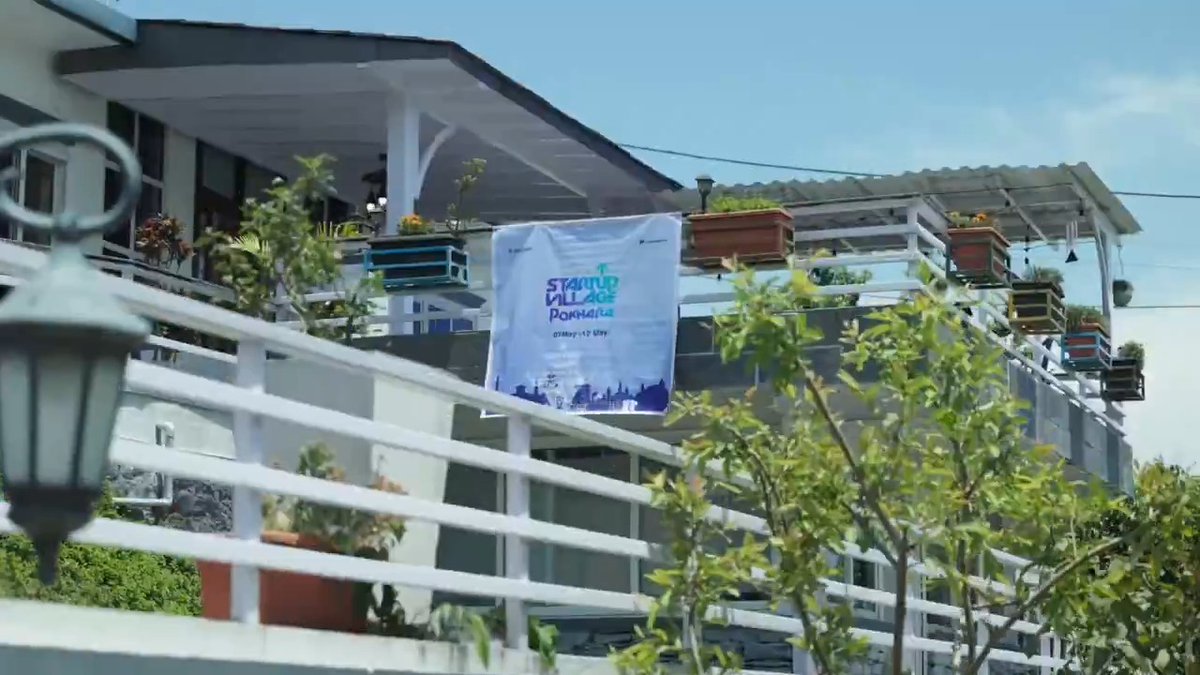 The image shows a beautiful building with a balcony overlooking the surrounding area. The building has a sign that reads "Startup Village" and features a banner hanging from its side, displaying the words "Startup Village". There are several potted plants on the balcony, adding greenery to the scene. A street lamp can be seen in the background, providing illumination for the area.