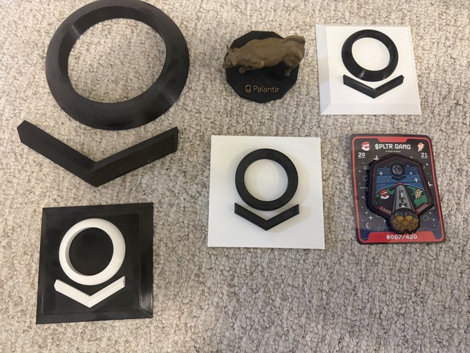 The image shows an assortment of $PLTR (Palantir) memorabilia laid out on a carpet, including a 3D-printed bull figurine on a Palantir-branded base, a large black ring/circle logo piece, a black chevron piece, and several Palantir logo stickers (both printed and 3D-printed), alongside a "Split Gang" trading card numbered #087/420, matching the caption's description of early 2021 Palantir investing memorabilia being offered up for free to followers.