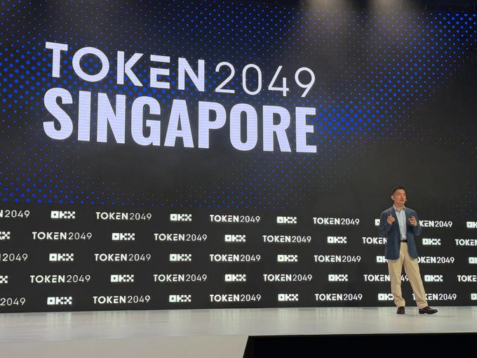 A man in a navy blazer and khaki pants speaks on stage in front of a large backdrop displaying "TOKEN 2049 SINGAPORE" with repeating logos.