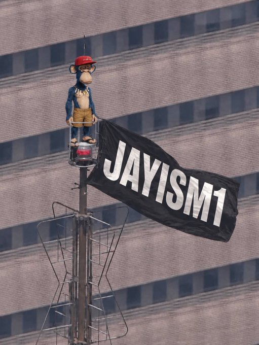 A monkey figurine wearing a red cap mounted on top of a pole with a black flag reading 'JAYISM1' against a building backdrop.