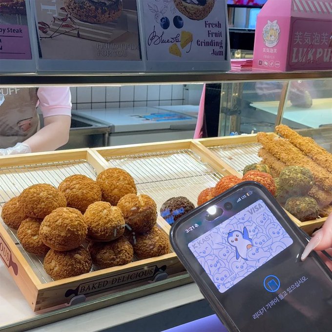 A phone displaying a 'TKAST' Visa card with a blue character design is held up near a bakery display case filled with breaded fried food items, with Korean text on the screen and signage.