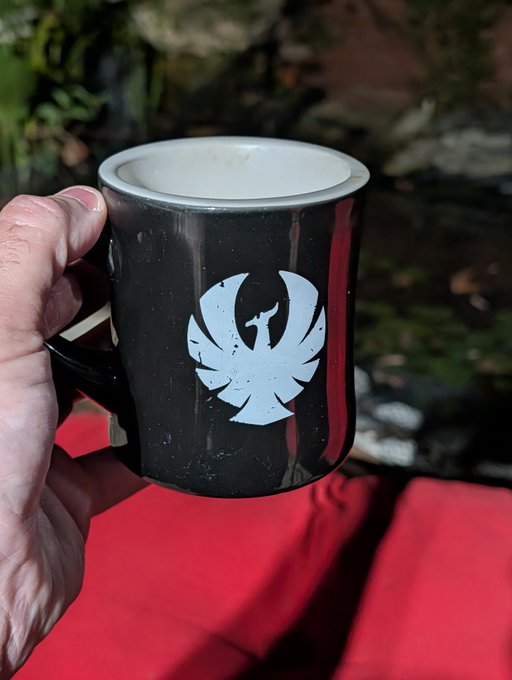 A hand holding a black enamel camping mug featuring a white stylized phoenix or bird logo, set against a backdrop of red fabric and outdoor greenery.