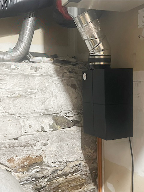 A black rectangular ventilation unit is mounted on a white wall against exposed stone, connected to silver flexible ductwork near the ceiling.