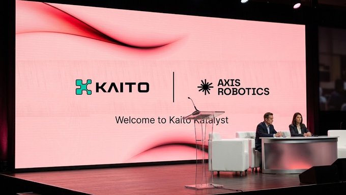 Two people are seated at a table on stage during the "Kaito Katalyst" launch event, with a large screen behind them displaying the KAITO and Axis Robotics logos alongside the welcome message. The image illustrates the announcement of Axis Robotics being the first token-based Katalyst campaign on the Kaito platform.