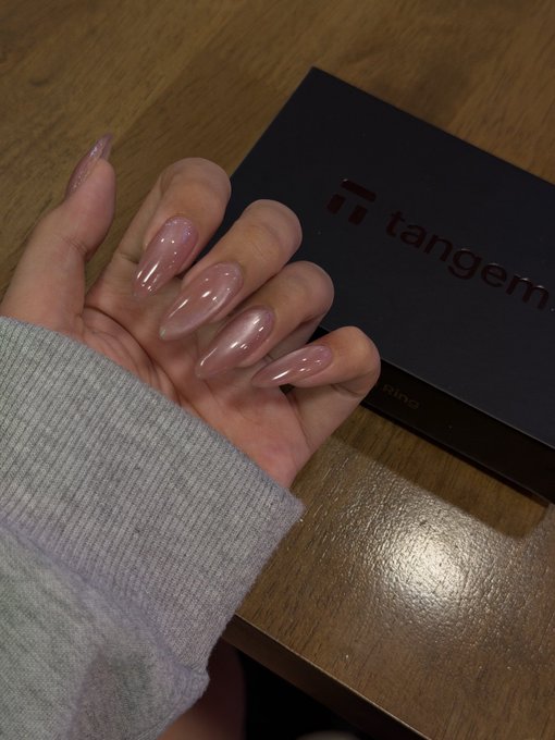 A hand showing off a fresh set of long, almond-shaped, glossy nude-pink acrylic nails, photographed over a 'tangem' branded card box on a wooden surface.