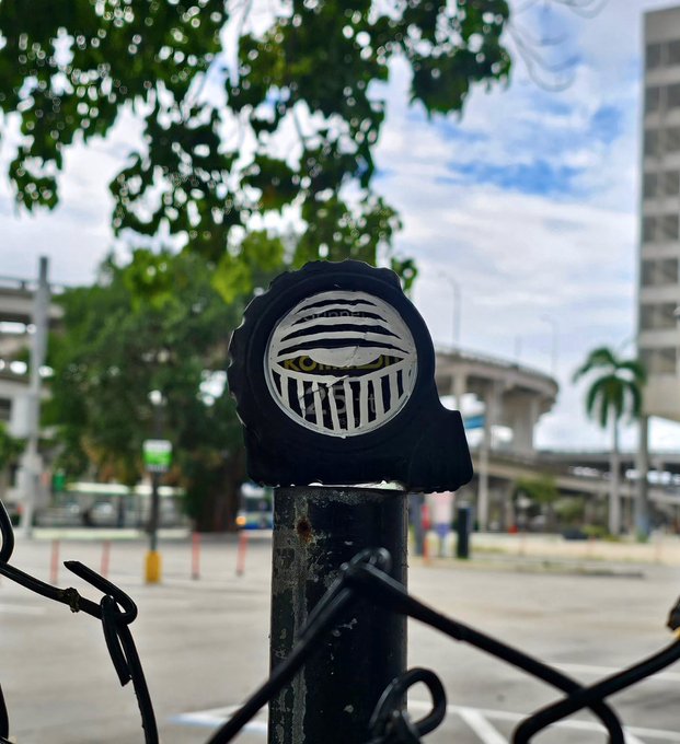 A street sign featuring a stylized grim reaper/helmet logo on a black post, with a blurred urban street scene and palm trees in the background.