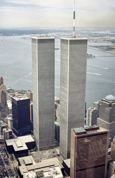 Aerial photograph of the Twin Towers of the World Trade Center in New York City, showing the two identical skyscrapers standing prominently with surrounding buildings and the water in the background.