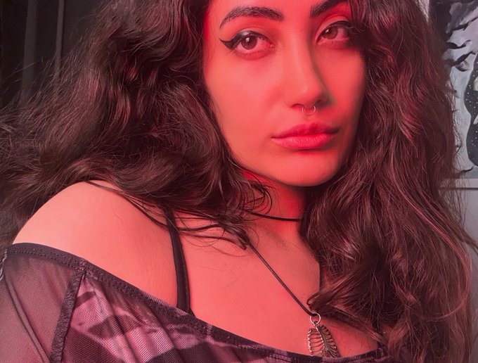 A close-up portrait of a young woman with long dark wavy hair, illuminated by red lighting, wearing a black mesh top and a pendant necklace, with a nose piercing.