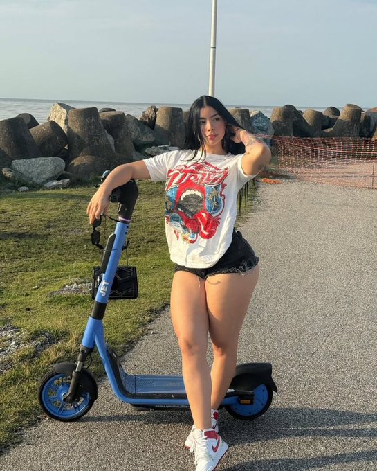 A young woman posing outdoors next to a blue and black electric scooter, wearing a graphic white t-shirt and black shorts, with a rocky shoreline and orange safety fencing visible in the background.