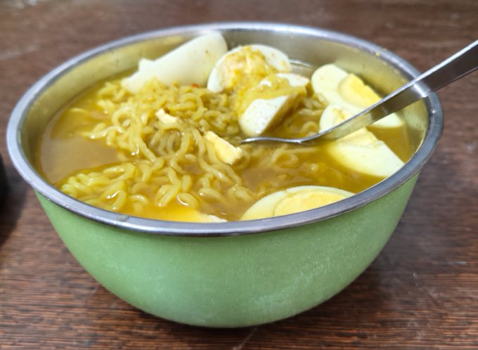 A green metal bowl with a stainless steel rim contains ramen noodles in a yellow broth, topped with sliced boiled eggs, with a spoon resting inside.
