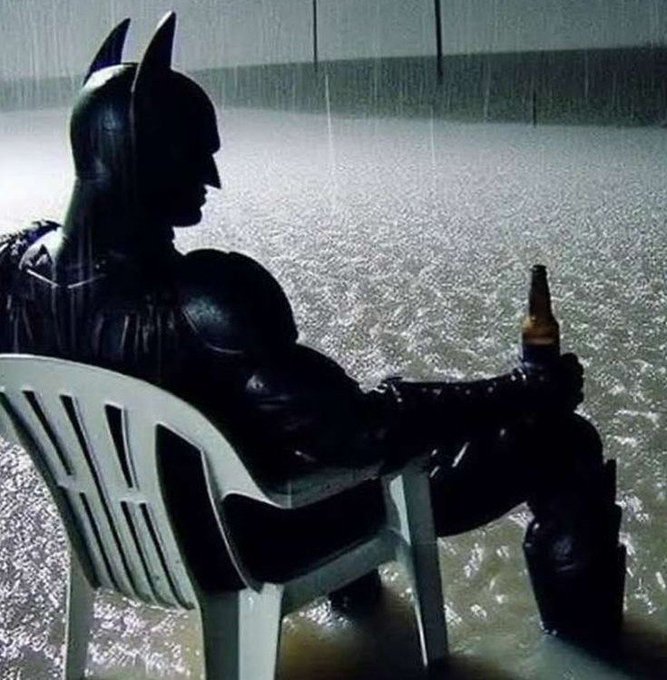 A person in a black Batman costume, sitting in a white plastic chair while holding a beer bottle, against a glittering silver background.