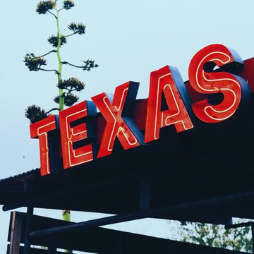 A bright red neon "TEXAS" sign glows against a clear blue sky, with a leafy plant visible in the background. The image evokes the state being referenced in the post about Texas lawmakers considering a ban on crypto kiosks following reported Bitcoin ATM scams.