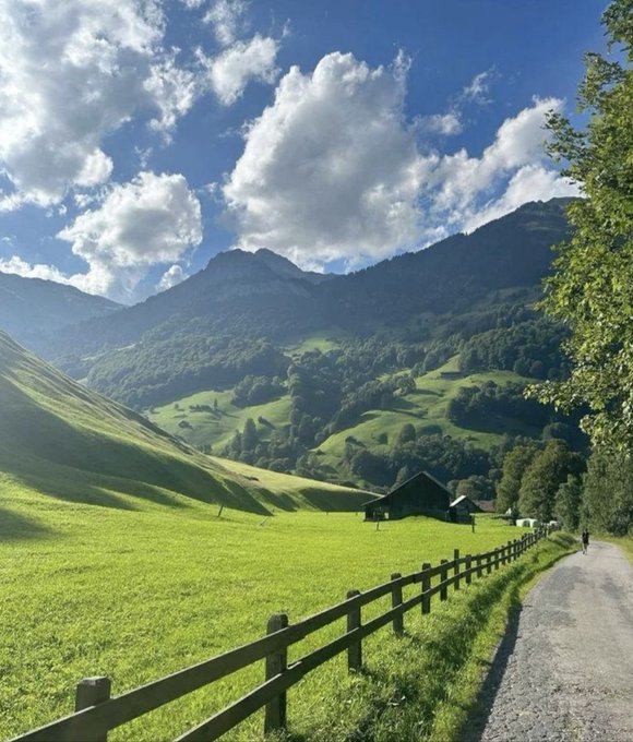 A scenic mountain landscape with lush green rolling hills, a wooden cabin nestled in the valley, a wooden fence alongside a paved path, and dramatic clouds against a blue sky.