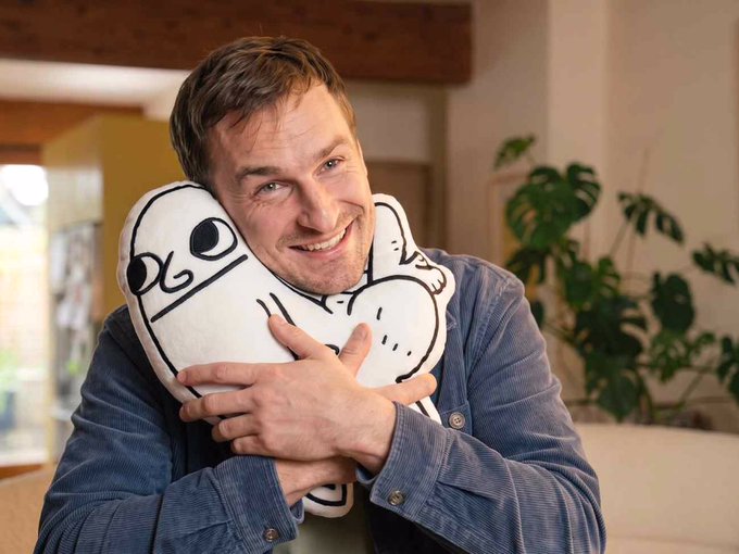 A smiling man with short brown hair hugging a white pillow with a black cartoon face design, sitting indoors with a plant in the background.