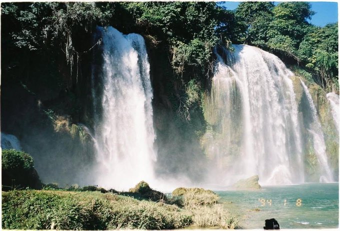 A scenic photograph of a large, multi-tiered waterfall surrounded by lush green trees, with a timestamp of '94 1 8' visible in the lower right.
