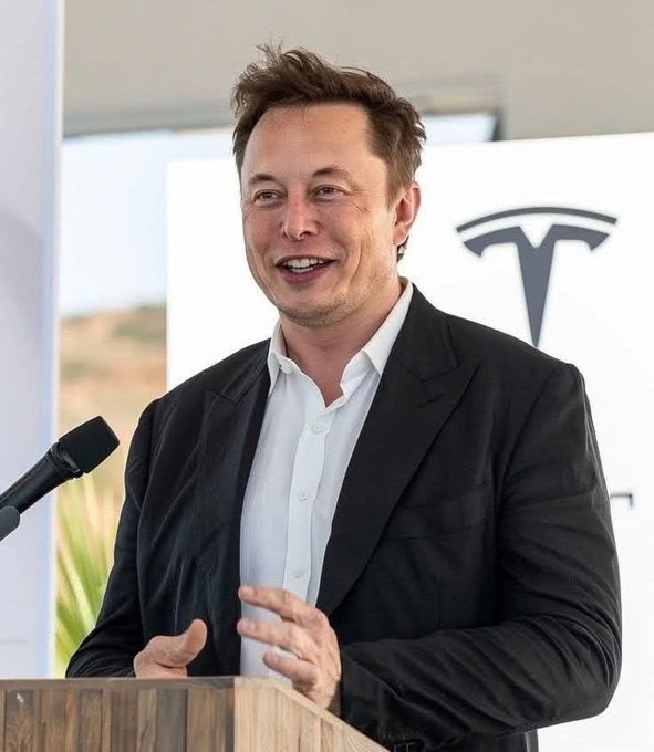Elon Musk smiles while speaking at a wooden podium with a microphone, wearing a black blazer over a white shirt, with a Tesla logo visible on a sign behind him.