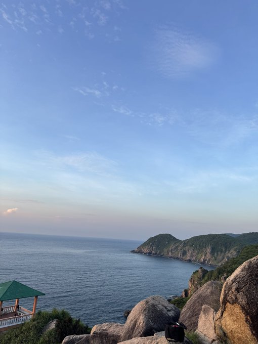 A serene coastal view at golden hour, with a traditional pavilion perched on rocky cliffs overlooking the calm sea and distant green islands. The peaceful seaside scenery suggests a perfect weekend escape, encouraging friends to swap their X/Twitter sessions for some fresh air and ocean views.