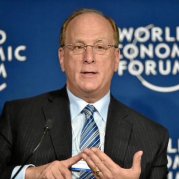 A man in a dark suit and blue striped tie speaks with his hands clasped, standing before a blue backdrop with the 'World Economic Forum' logo.