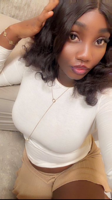 A selfie of a young woman with dark hair wearing a white long-sleeved top and tan skirt, accessorized with delicate gold necklaces and a bracelet.
