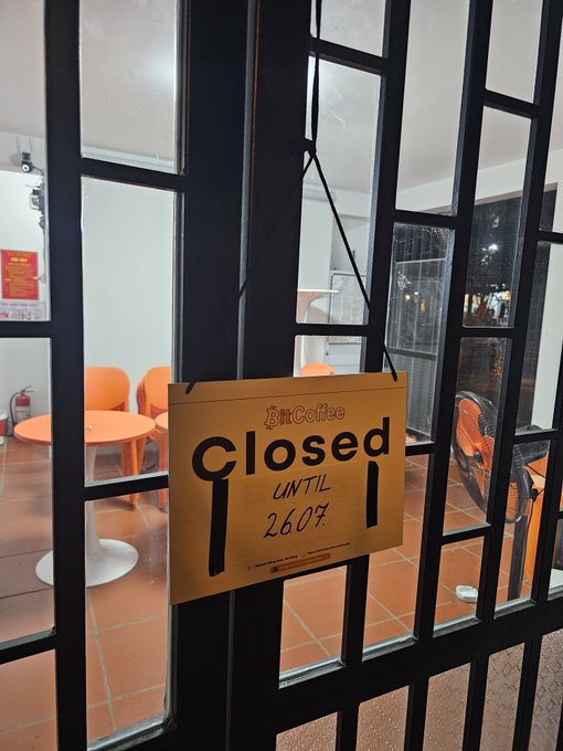 A handwritten 'Closed until 26.07' sign hanging on the glass door of 'BnCoffee', showing an empty interior with orange chairs.