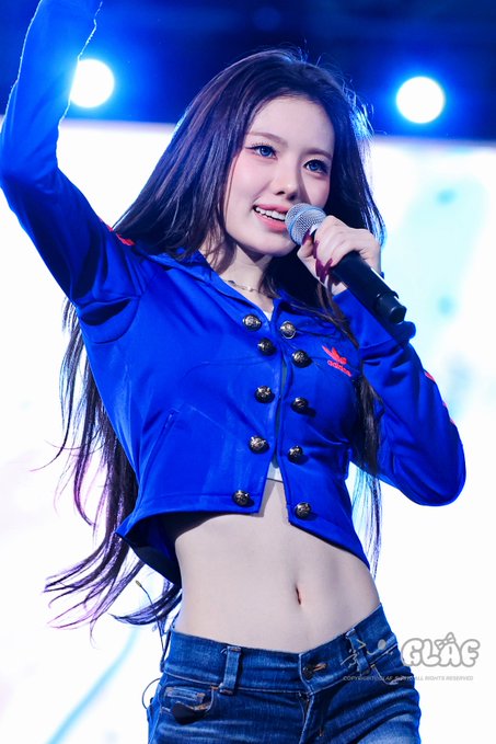 A performer with long dark hair wearing a blue cropped jacket with gold buttons sings into a microphone on a brightly lit stage.