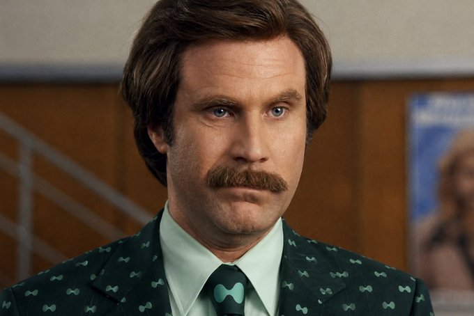 A man with brown hair and a mustache wearing a dark suit with light green bowtie patterns and a matching bowtie, looking directly at the camera.