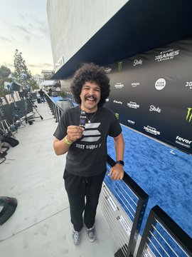 A man with curly hair and a mustache smiles while wearing a black t-shirt and white sneakers, posing on a blue carpet in front of a step-and-repeat banner featuring various logos.