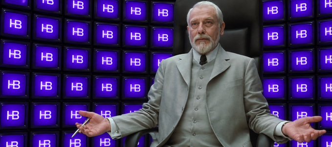 An older bearded man in a gray suit stands with arms outstretched in front of a wall of blue television screens displaying the 'HB' logo.