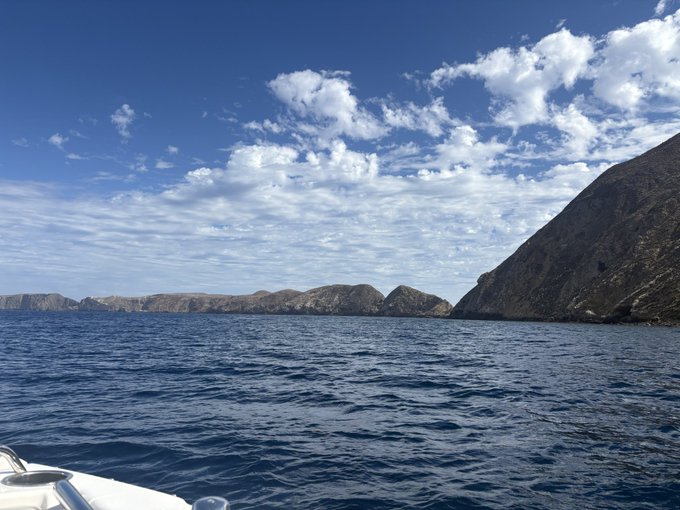 A scenic ocean view from a boat showing rocky cliffs and distant mountains under a blue sky with scattered clouds.