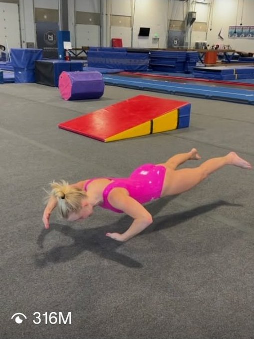 The image features a young girl wearing a pink swimsuit, lying on her stomach with her arms extended outwards as she performs a gymnastics exercise on a mat. The setting appears to be an indoor gymnasium or sports facility, which is filled with various equipment and objects such as a ball, a ramp, and other gymnastic apparatus. A computer monitor can also be seen in the background.