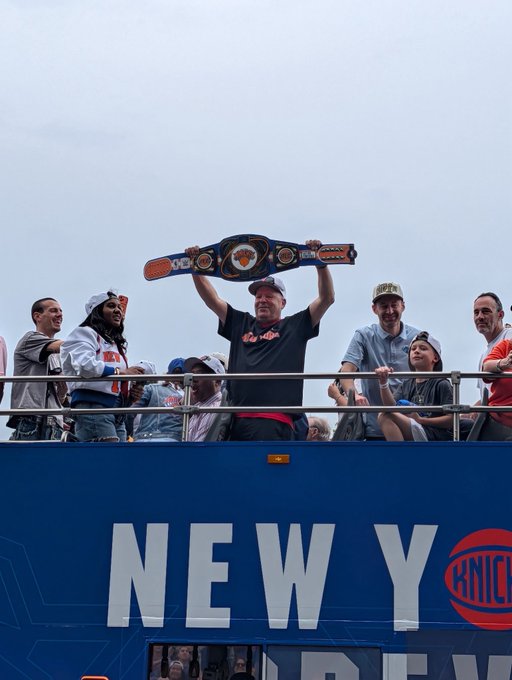 In the image, a man wearing a black shirt is holding up a large weight above his head while standing on top of a bus. The bus has an advertisement for the New York Knicks on its side. There are several people around him who appear to be cheering or clapping in excitement. A crowd can also be seen in the background, indicating that this event might be taking place at a sports venue or a similar public space.