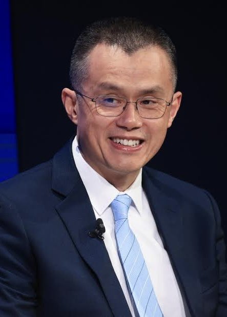 A smiling man in a dark suit, light blue striped tie, and glasses appears on stage with a lapel microphone attached to his jacket, likely Changpeng Zhao, founder of Binance, the cryptocurrency exchange mentioned in the accompanying post about limiting commodity perpetual futures trading to traditional 24/5 market hours.