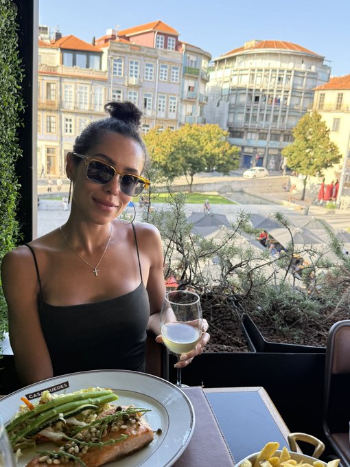 A woman wearing sunglasses and a black top holds a glass of white wine while seated at an outdoor restaurant table with a plate of food including asparagus and salmon.