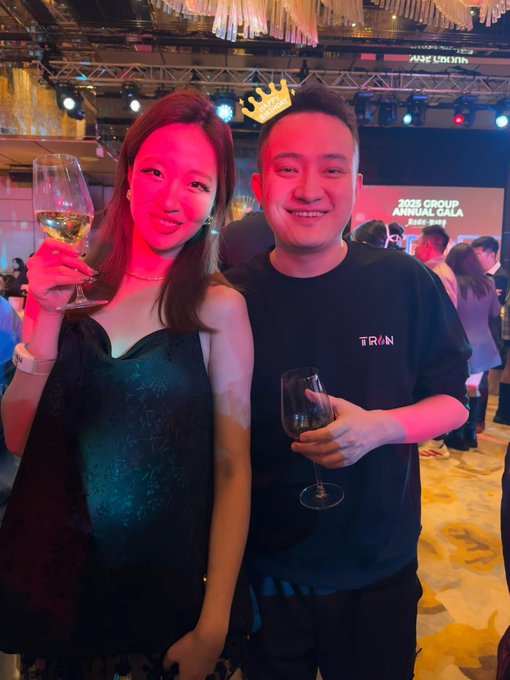 A man and a woman pose for a photo at a gala event; the woman wears a black strapless dress and holds a glass of white wine, while the man in a 'TRON' t-shirt holds a red wine glass and has a gold crown graphic edited above his head.