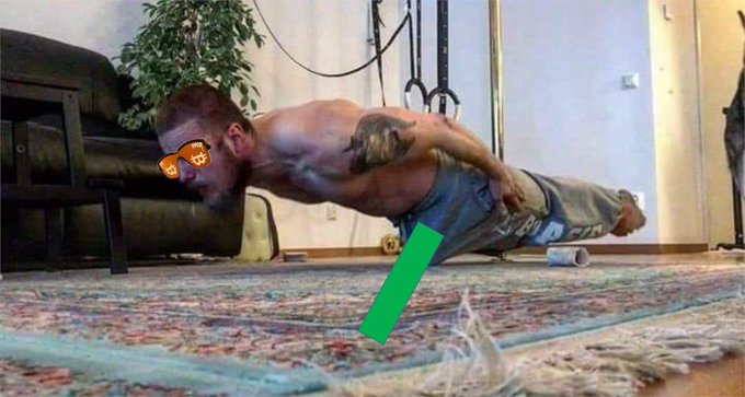 A shirtless man with a beard and tattoos performing a planche exercise on a rug in a living room, wearing cargo shorts and sunglasses, with a green rectangle overlaid near his hands.