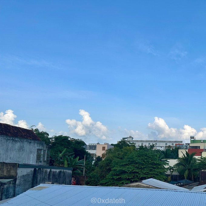 A bright blue sky with scattered clouds rises above a cityscape of rooftops and lush green trees, with a watermark reading '@0xdateth' at the bottom.