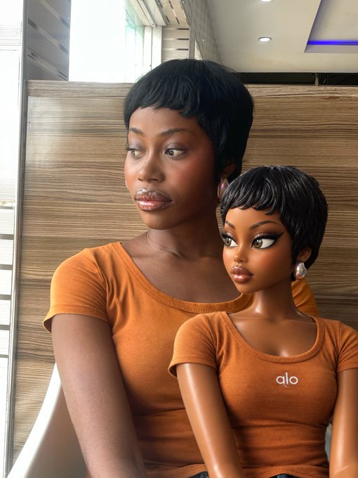 A woman in an orange "alo" t-shirt poses beside a digitally rendered doll designed to resemble her, both featuring short black pixie cuts.