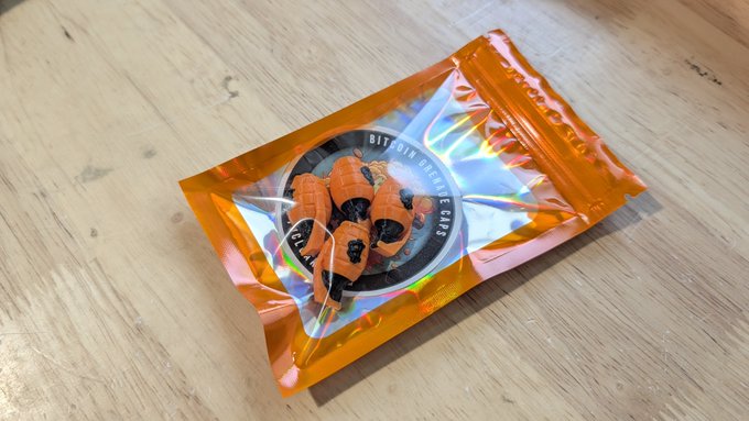 An orange metallic Mylar package containing orange and black striped items, possibly candy or small toys, sits on a wooden floor, with text visible on the inner label including '24128'.