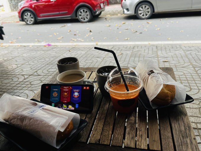 A wooden table outside a cafe set with iced coffee in plastic cups, paper-wrapped sandwiches/burgers, and a smartphone playing a mobile game, with parked cars visible on the street behind.