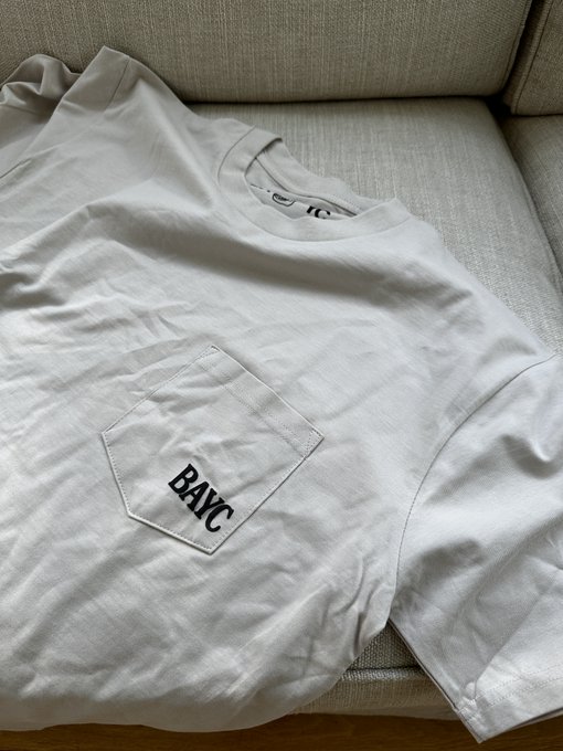 A plain light gray t-shirt laid on a beige couch, featuring a chest pocket with the embroidered text 'BAYC' (Bored Ape Yacht Club).