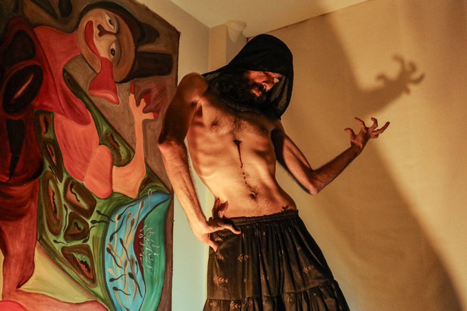 A shirtless man with a beard, wearing a black hood and a long skirt-like garment, poses with claw-like hand gestures in front of an abstract colorful painting.