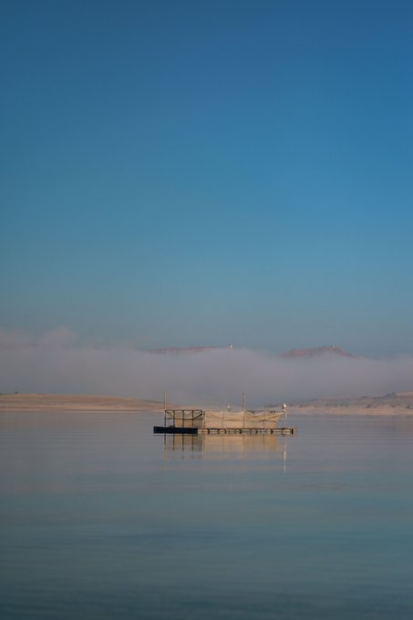 A serene lake scene with a small wooden dock or platform in the center, surrounded by mist-covered hills under a clear blue sky.